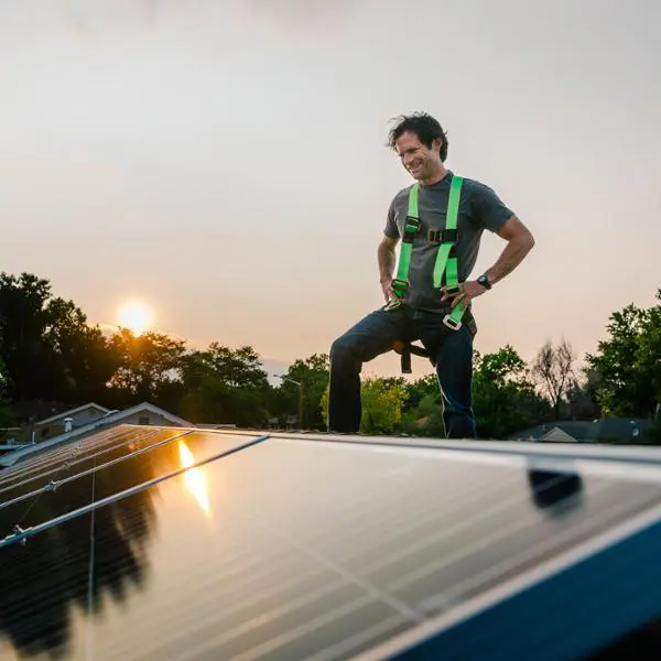 Person i säkerhetssele står på ett tak med solpaneler i solnedgången. Hållbar energiinstallation