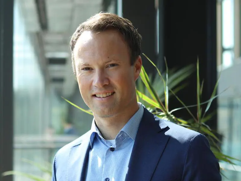 Man i blå kostym och skjorta, leende, stående inomhus med växter i bakgrunden