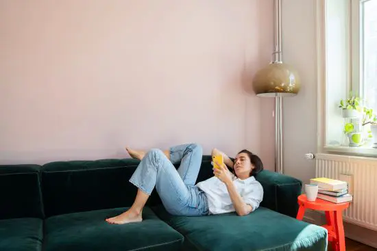 Kvinna avslappnad läsande på en mörkgrön soffa i ett ljust rum med en bok i handen, ett litet rött bord bredvid med böcker och en vit kopp